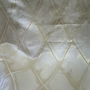 May include: Close-up of a cream-colored fabric with a diamond pattern. The diamonds are outlined with a lighter, metallic gold color, creating a textured effect. The fabric has a subtle sheen.