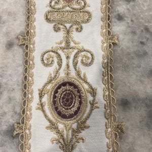 May include: A white fabric trim featuring an intricate embroidered pattern in metallic gold thread. The design includes symmetrical scrollwork, floral motifs, and circular accents in deep burgundy. The edges are finished with a scalloped lace border.