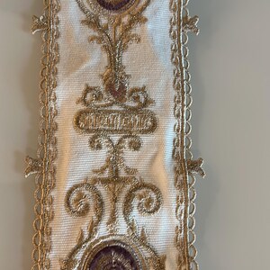 May include: A white fabric trim featuring intricate gold metallic embroidery and circular purple accents. The design includes symmetrical scrollwork and floral-inspired motifs, finished with a delicate scalloped lace edge along both sides.