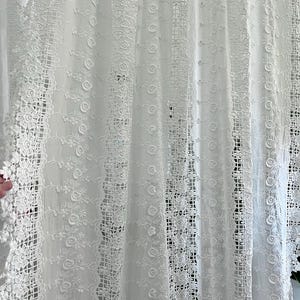 May include: White lace curtain with floral and geometric patterns. The curtain features intricate details, including roses and openwork designs. The fabric appears to be delicate and airy, creating a soft, romantic aesthetic.