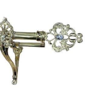 May include: A gold-toned curtain rod with ornate, floral-inspired detailing. The rod features a decorative end with a light blue, oval-shaped gem. The support bracket has a curved design with leaf-like accents, adding a touch of elegance.