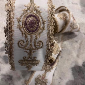 May include: A roll of white ribbon featuring intricate gold metallic embroidery and a central purple oval motif. The ribbon edges are finished with a decorative scalloped gold lace trim, creating a textured and detailed appearance throughout the length.