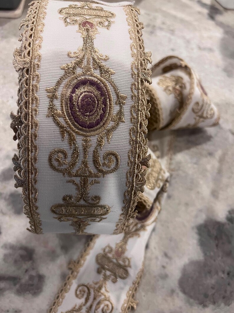 May include: A roll of white ribbon featuring intricate gold metallic embroidery and a central purple oval motif. The ribbon edges are finished with a decorative scalloped gold lace trim, creating a textured and detailed appearance throughout the length.