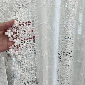 May include: Close-up of white lace fabric with a floral design. The lace features detailed, openwork patterns of flowers and leaves, creating a delicate and intricate texture. The fabric is likely used for curtains or decorative purposes.