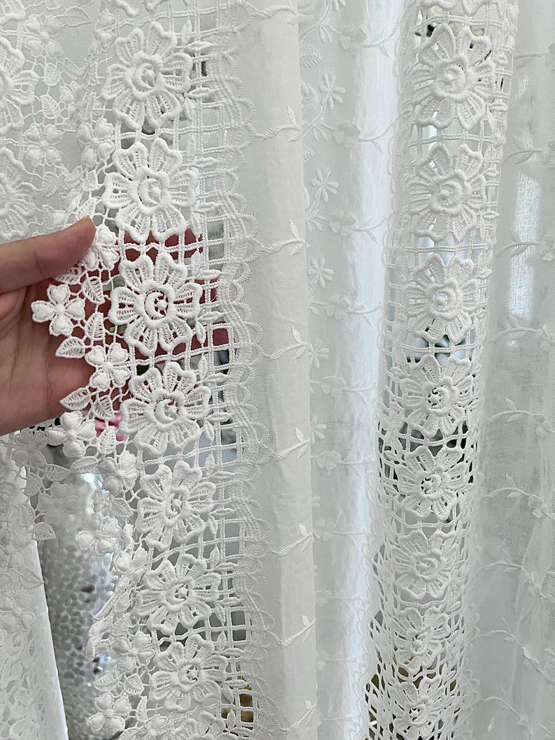 May include: Close-up of white lace fabric with a floral design. The lace features detailed, openwork patterns of flowers and leaves, creating a delicate and intricate texture. The fabric is likely used for curtains or decorative purposes.