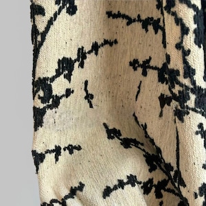May include: Close-up of a beige fabric with a black floral pattern. The design features stylized branches and leaves, creating a textured, organic look. The fabric appears to be woven, with a slightly rough surface.