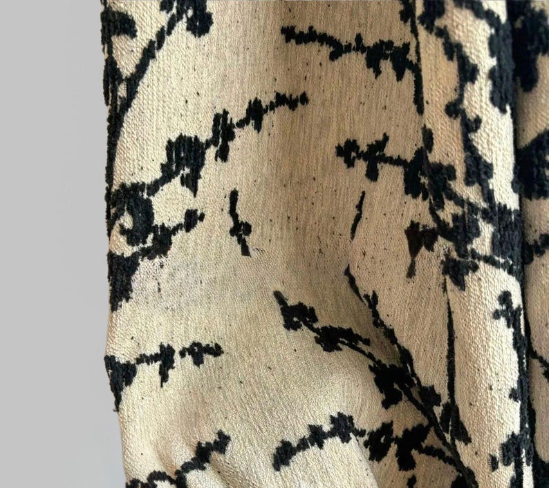 May include: Close-up of a beige fabric with a black floral pattern. The design features stylized branches and leaves, creating a textured, organic look. The fabric appears to be woven, with a slightly rough surface.