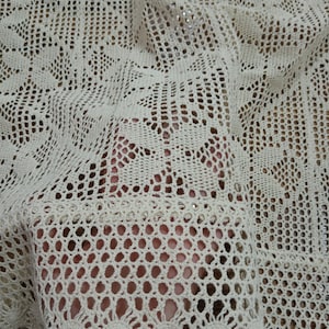 以下が含まれることがあります： オフホワイトの、複雑な透かし模様の入ったかぎ針編みの生地のクローズアップ。繊細な織物は花柄と幾何学模様を特徴とし、質感と風通しの良い外観を作り出しています。生地はドレープされており、その柔軟性とドレープ性を際立たせています。