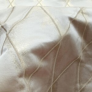 May include: Close-up of a fabric with a diamond pattern. The fabric has a shimmering, textured surface with a light beige color. The diamond pattern is created by thin, light gold lines.