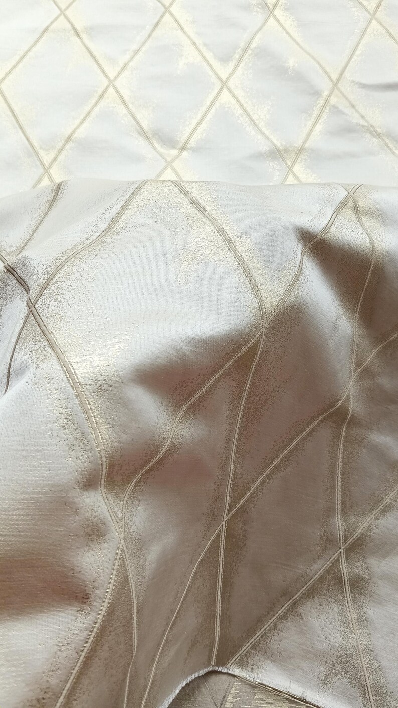 May include: Close-up of a fabric with a diamond pattern. The fabric has a shimmering, textured surface with a light beige color. The diamond pattern is created by thin, light gold lines.