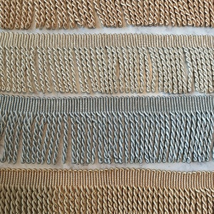 May include: Close-up of four rows of decorative fringe trim in various metallic colors. The colors include gold, silver, and bronze. Each row features a twisted rope design, adding texture and visual interest. Ideal for home decor and craft projects.