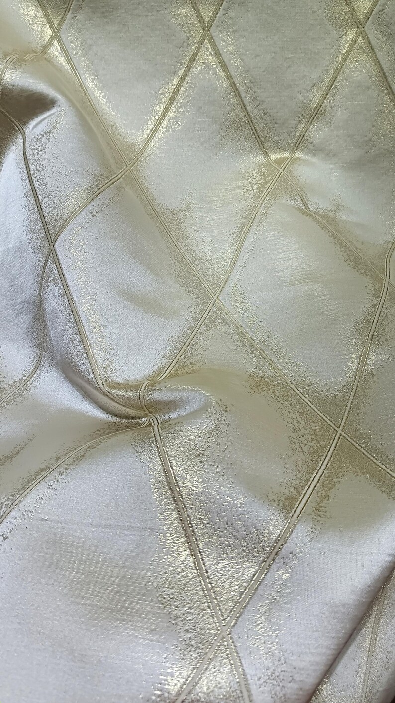 May include: Close-up of a shimmering fabric with a diamond pattern. The fabric has a soft, textured appearance with a light cream base and gold accents. The diamond grid is defined by gold lines.