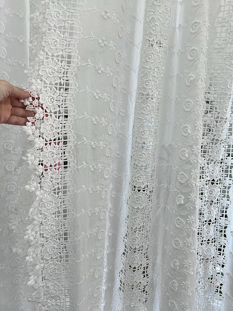 May include: White lace curtain with floral and geometric patterns. The curtain features intricate embroidered details, including flowers and openwork designs. The delicate fabric is semi-sheer, allowing light to filter through.
