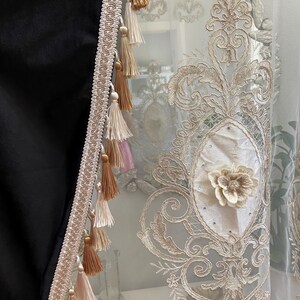 Pu&ograve; includere: Primo piano di una tenda decorativa con ricami intricati e un pannello nero. La tenda presenta un motivo floreale color crema, impreziosito da un fiore 3D e nappe nei toni del beige e del marrone. Il bordo &egrave; beige chiaro.