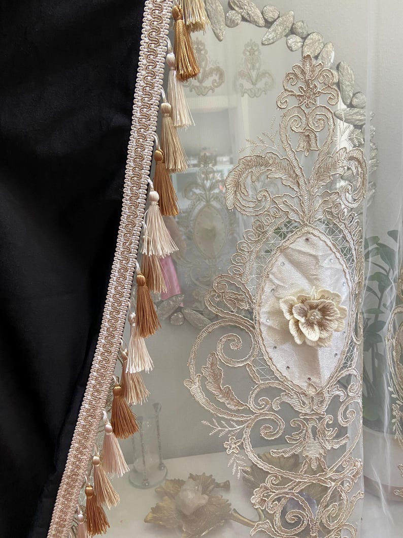 Pu&ograve; includere: Primo piano di una tenda decorativa con ricami intricati e un pannello nero. La tenda presenta un motivo floreale color crema, impreziosito da un fiore 3D e nappe nei toni del beige e del marrone. Il bordo &egrave; beige chiaro.