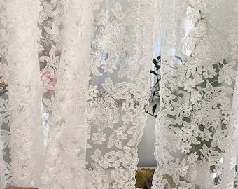 May include: Sheer white lace curtains with a delicate floral pattern. The curtains are semi-transparent, allowing light to filter through. The intricate design features various flowers and leaves, creating an elegant and airy aesthetic. The fabric appears soft and flowing.
