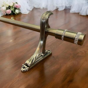 May include: A bronze-colored curtain rod with decorative end caps and a matching bracket. The end cap features two rows of sparkling rhinestones. The bracket has a stylized leaf design. The rod is displayed on a wood surface.