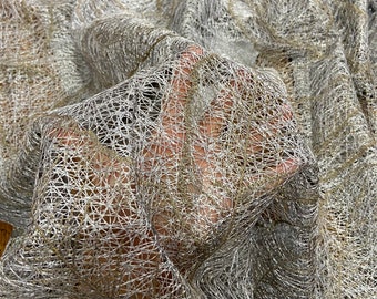 Pode incluir: Close-up de um tecido transparente tecido com uma trama delicada e aberta. O tecido é principalmente prateado com sutis fios dourados tecidos, criando um efeito brilhante. A textura é visível, com uma aparência ligeiramente enrugada. O tecido está drapeado.