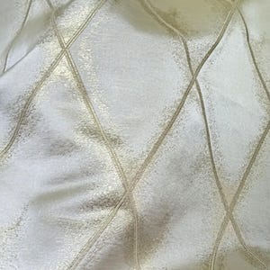 May include: Close-up of a cream-colored fabric with a diamond pattern. The pattern is created by gold-colored lines that intersect, forming a grid-like design. The fabric has a subtle sheen, suggesting a luxurious texture.