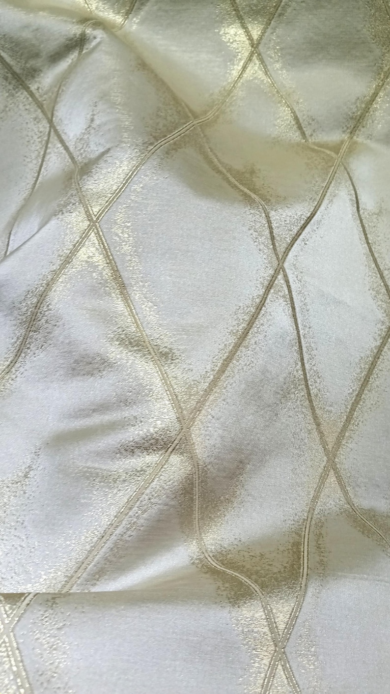 May include: Close-up of a cream-colored fabric with a diamond pattern. The pattern is created by gold-colored lines that intersect, forming a grid-like design. The fabric has a subtle sheen, suggesting a luxurious texture.