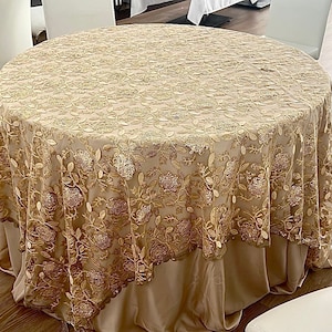 May include: A round table covered with a gold-colored tablecloth. The tablecloth features an intricate floral embroidery design. The table is set in a room with white chairs and a wooden floor. The tablecloth has a skirt.