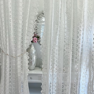 May include: White lace curtains with intricate floral and geometric patterns. The curtains frame a glimpse of a room with a decorative mirror, a silver vase, and a white dresser. The overall aesthetic is delicate and romantic.