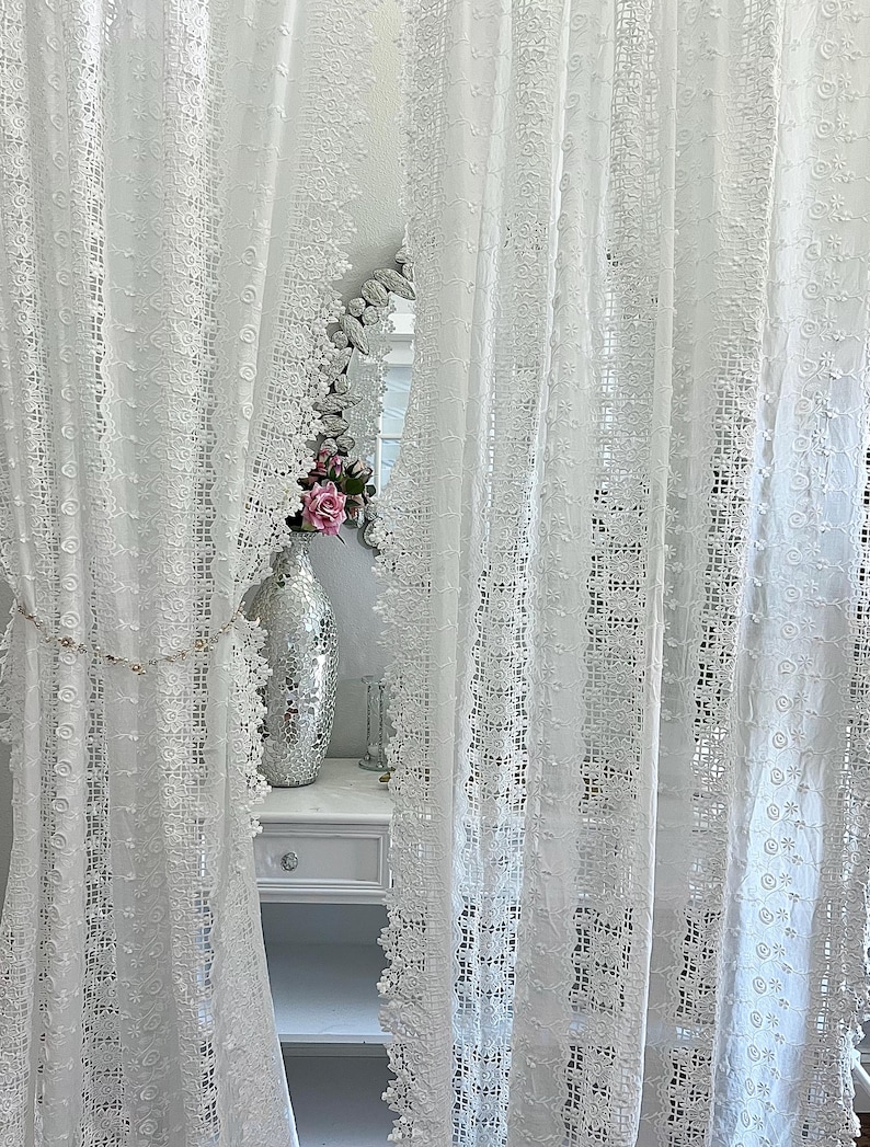 May include: White lace curtains with intricate floral and geometric patterns. The curtains frame a glimpse of a room with a decorative mirror, a silver vase, and a white dresser. The overall aesthetic is delicate and romantic.