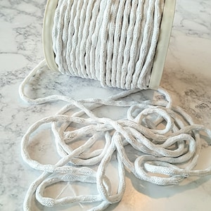 May include: A spool of white rope with a braided texture. The rope is coiled on a white plastic spool, with some rope unraveled and lying on a light-colored surface. The rope appears to be made of a soft, flexible material.