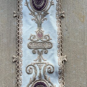 May include: A white fabric trim featuring an intricate embroidered pattern in metallic gold thread. The design includes repeating circular motifs with deep purple centers, flanked by decorative scrollwork and finished with a scalloped lace edge.