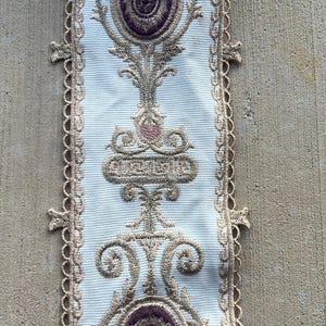 May include: A white fabric trim featuring intricate gold embroidery and deep purple oval accents. The design includes symmetrical scrollwork and floral-inspired motifs. The edges are finished with a delicate, scalloped gold lace border.