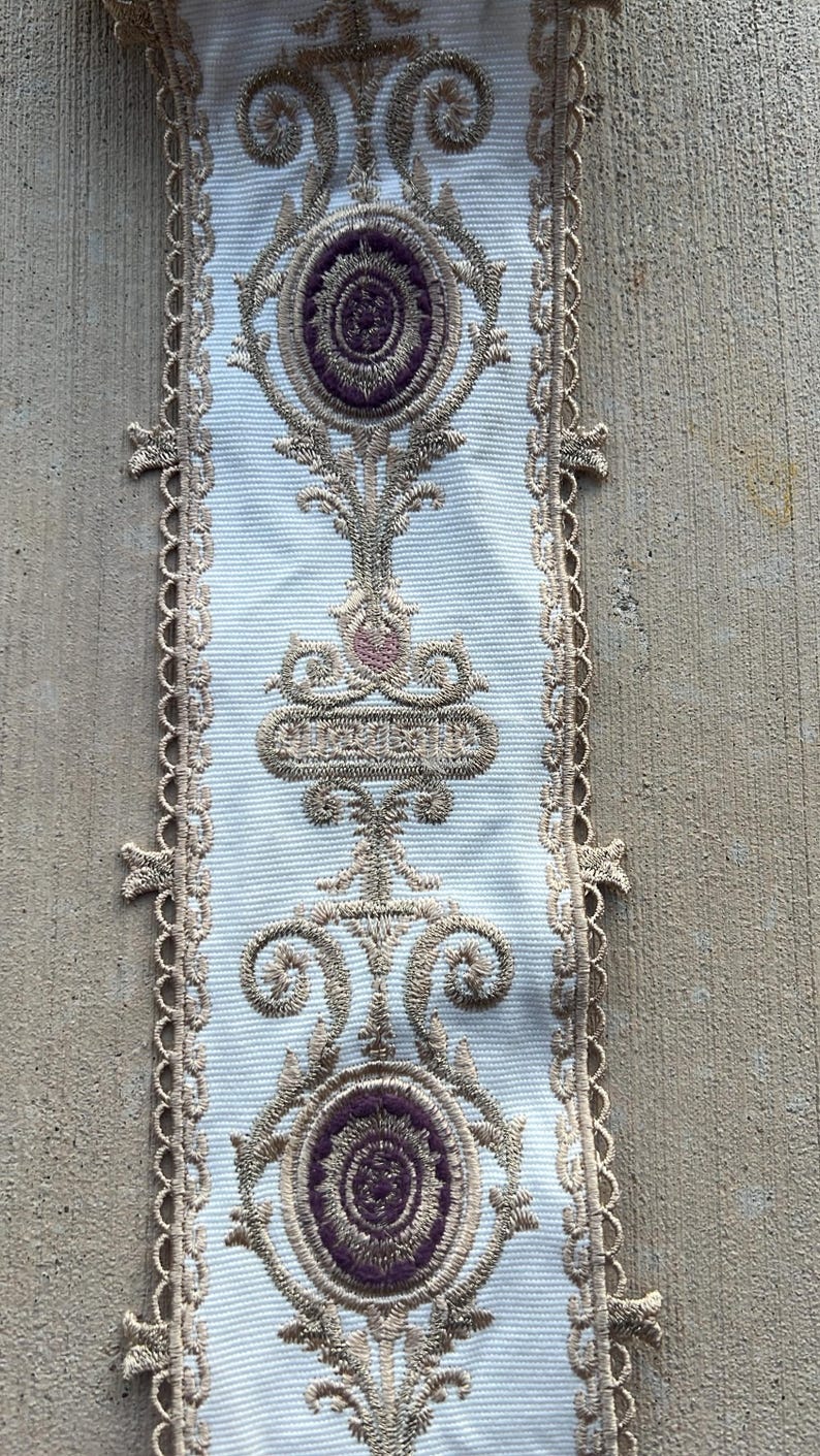 May include: A white fabric trim featuring intricate gold embroidery and deep purple oval accents. The design includes symmetrical scrollwork and floral-inspired motifs. The edges are finished with a delicate, scalloped gold lace border.