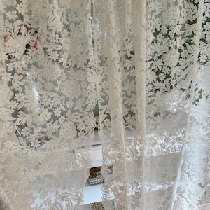 May include: Cream-coloured lace curtains with a floral design. The curtains are semi-transparent, allowing light to pass through. The detailed pattern features different flowers and leaves, creating a delicate and elegant appearance. The curtains are hanging in front of a window.