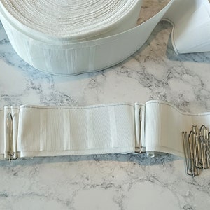 May include: A roll of white curtain heading tape with vertical ribbed texture. Several metal curtain hooks are next to a section of the tape with hooks attached. The tape is on a marble-patterned surface.
