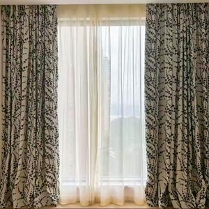 May include: A set of window curtains featuring a botanical design. The curtains are a light beige with a black leaf pattern. Sheer white curtains are in the centre. The curtains are hung in front of a window.