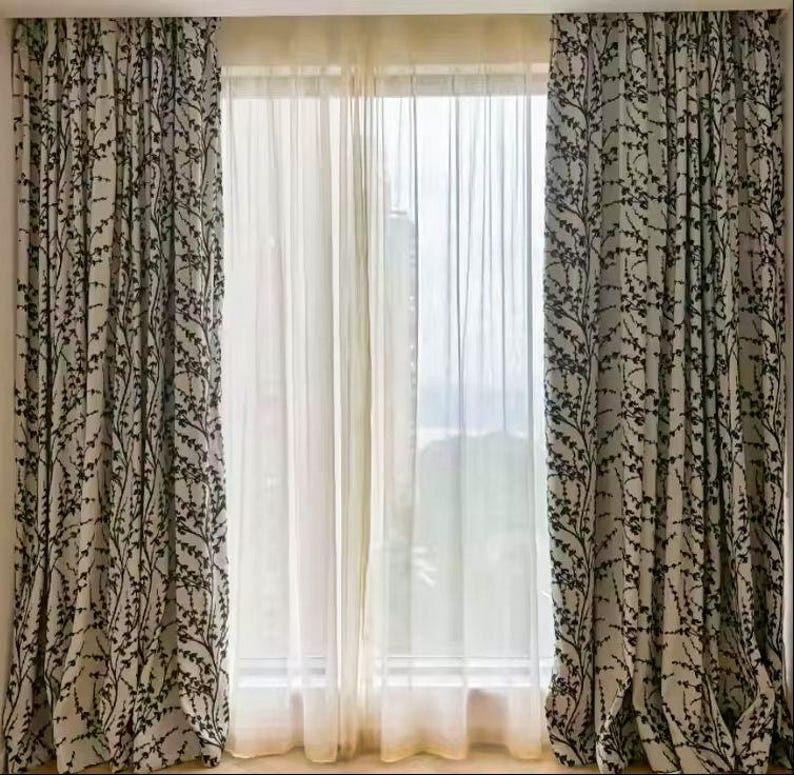 May include: A set of window curtains featuring a botanical design. The curtains are a light beige with a black leaf pattern. Sheer white curtains are in the centre. The curtains are hung in front of a window.