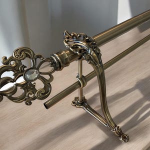 May include: Ornate antique brass curtain rod with decorative finials. The rod features a scrolling floral design with a central clear crystal accent. The support brackets and rod have a vintage aesthetic, suitable for classic interior design.