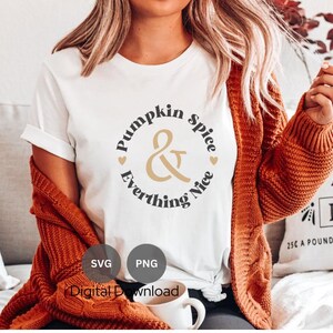 Peut inclure: Un t-shirt blanc avec le texte "Pumpkin Spice & Everything Nice" dans un cercle de couleur or et noir. Le t-shirt est porté par une personne aux cheveux longs bruns et un cardigan marron.