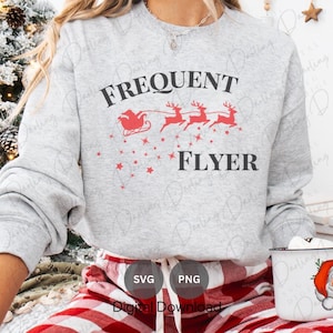 以下が含まれることがあります： 「Frequent Flyer」という文字と、トナカイに引かれたそりに乗ったサンタクロースのグラフィックが描かれたグレーのスウェットシャツ。赤と白のチェック柄のパンツを履いた人が着用しています。