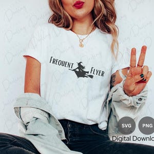 以下が含まれることがあります： 「Frequent Flyer」という文字と、ほうきに乗って飛んでいる魔女のシルエットが描かれた白いTシャツ。文字は黒で、魔女は灰色。長い茶色の髪の女性が着ています。