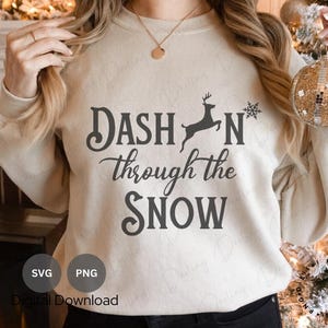 May include: A cream-colored sweatshirt with the text "Dash'n through the snow" and a silhouette of a reindeer with antlers. The text is in a black cursive font. The sweatshirt is on a person wearing black pants.