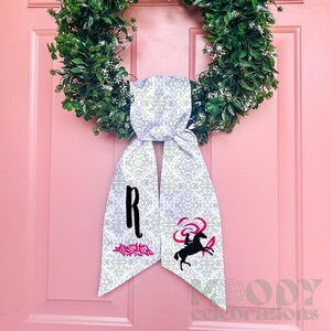 May include: A decorative door hanger featuring a green wreath and a white scarf with a floral pattern. The scarf has a large black letter "R", a pink floral design, and a black horse graphic with pink accents.