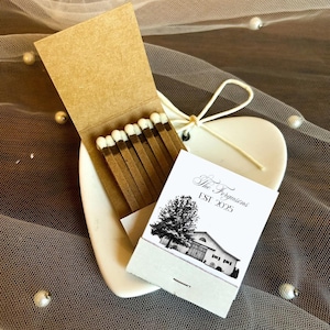 Puede incluir: Una caja de cerillas con cerillas marrones y una etiqueta impresa en blanco y negro. La etiqueta dice "The Fergusons EST 2025" y presenta un &aacute;rbol y una ilustraci&oacute;n de un edificio. La caja de cerillas est&aacute; sobre un plato blanco en forma de coraz&oacute;n.