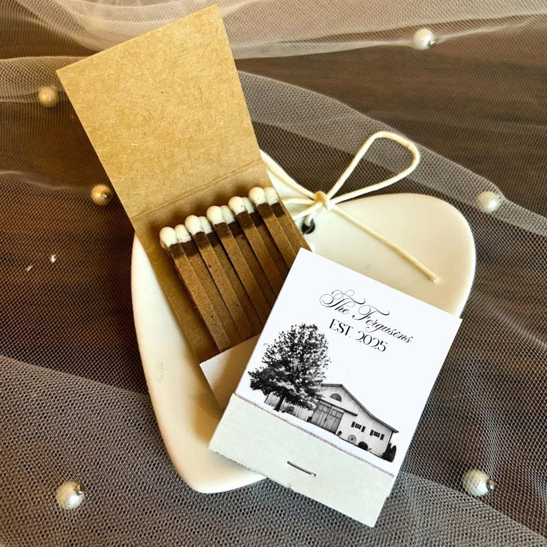 Puede incluir: Una caja de cerillas con cerillas marrones y una etiqueta impresa en blanco y negro. La etiqueta dice "The Fergusons EST 2025" y presenta un &aacute;rbol y una ilustraci&oacute;n de un edificio. La caja de cerillas est&aacute; sobre un plato blanco en forma de coraz&oacute;n.