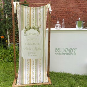 May include: A vertical fabric banner hanging on a wooden frame featuring a sage green bow graphic and text that reads Welcome to Jessica's Baby Shower Baby Taylor Coming July 2026, with vertical stripes in shades of green, yellow, and white.