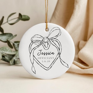 May include: A white ceramic ornament with a gold string for hanging. The ornament features a black line drawing of a heart-shaped tag with a bow and the name "Jessica", the word "OFFICIANT", and the date "5.9.26".