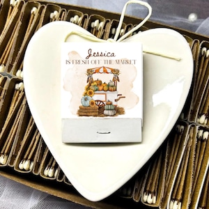 May include: A matchbox with a fall-themed illustration featuring pumpkins, sunflowers, and a market cart. The text on the matchbox reads "Jessica IS FRESH OFF THE MARKET." The matchbox is placed on a heart-shaped, off-white ceramic dish, surrounded by matchsticks.