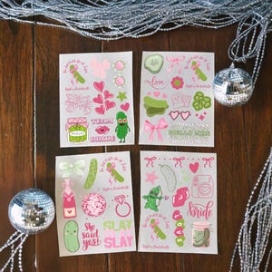 May include: Four sheets of stickers with a pickle theme, featuring pink and green designs. The stickers include hearts, bows, pickles, and text like "Bride", "Slay", and "She said yes!". Decorative disco balls and silver garland are in the background.