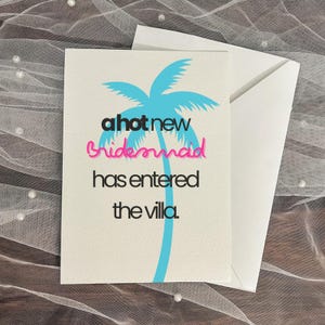 May include: A white card with the text "a hot new bridesmaid has entered the villa" in black, pink, and blue. A blue palm tree graphic is behind the text. A white envelope is behind the card.