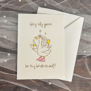 May include: A white greeting card with the text "Hey silly goose be my bridesmaid?" features a cartoon goose wearing a veil, pink shoes, and holding a bottle and a glass of yellow liquid. The card is paired with a white envelope, set against a textured background.