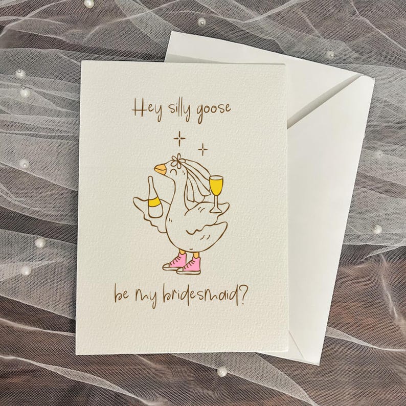 May include: A white greeting card with the text "Hey silly goose be my bridesmaid?" features a cartoon goose wearing a veil, pink shoes, and holding a bottle and a glass of yellow liquid. The card is paired with a white envelope, set against a textured background.